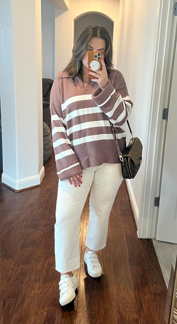 A person dressed in a striped sweater, white pants, and sneakers, taking a mirror selfie indoors. They’re holding a phone with a pop socket and sporting a stylish shoulder bag.