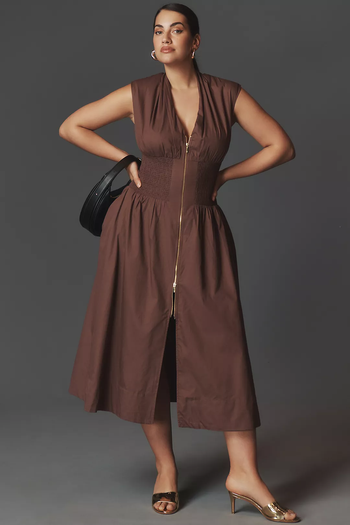 A model wearing a sleeveless, knee-length brown dress with a cinched waist and front zipper, holding a black handbag and paired with open-toe heels.