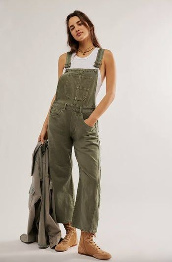 A person wearing olive-green overalls, a white tank top, brown boots, and a layered necklace, confidently posing while holding a matching olive-green jacket.