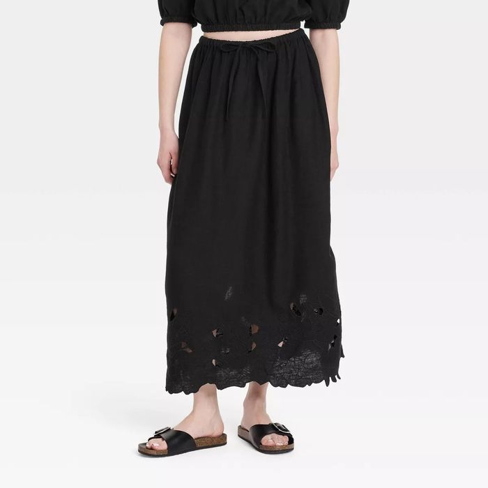 A person wearing a black maxi skirt featuring a floral cutout hem and black sandals, standing against a plain background with their face not visible