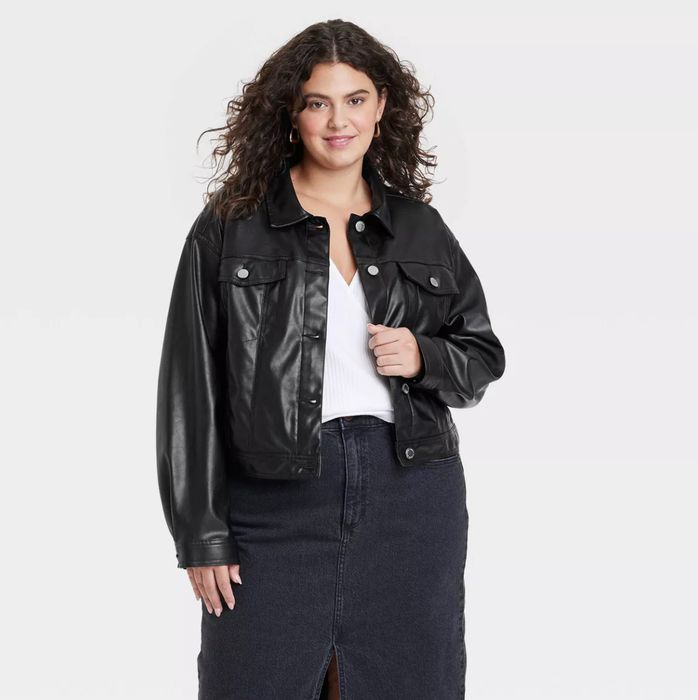A curly-haired model wearing a black leather jacket, a white top, and a dark skirt, smiling as she poses against a plain background