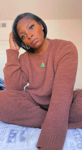 A reviewer sits on a bed, dressed in a warm brown knit sweater and matching pants.