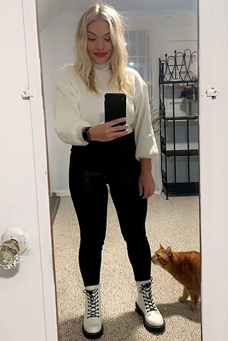 reviewer styling the white boots with black pants and a white sweater, posing next to their cat