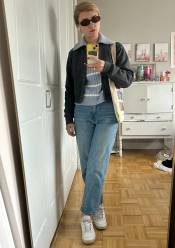 A person in a casual outfit featuring sunglasses, a striped sweater, jeans, sneakers, and a tote bag, taking a mirror selfie in a bedroom.