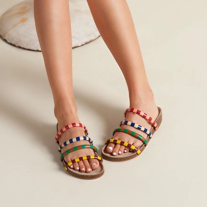 A person is sitting comfortably, sporting vibrant multi-colored sandals with straps, and a white dress adorned with lace trim, giving off a breezy, summertime vibe.