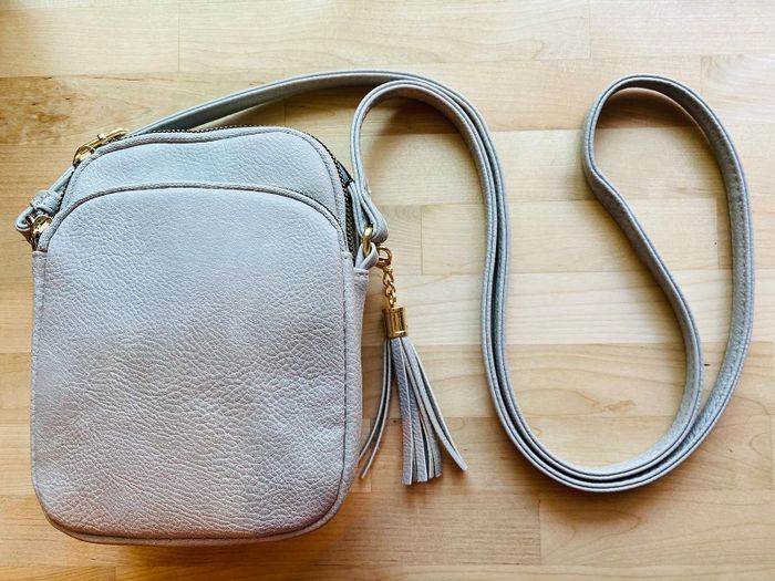reviewer’s light grey crossbody bag featuring a zippered compartment and tassel detail, displayed on a wooden surface