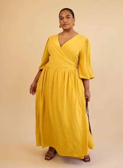 A model posing in a flowing, long-sleeved yellow dress with a wrap-style bodice and ankle-length hem, perfect for a shopping feature.