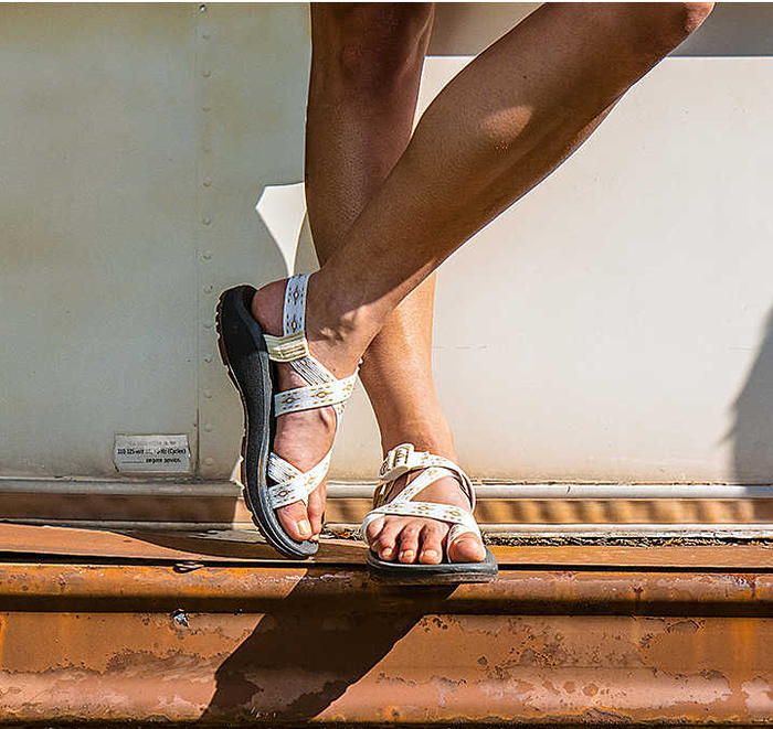 A person wearing white strappy sandals stepping on a rusty metal surface