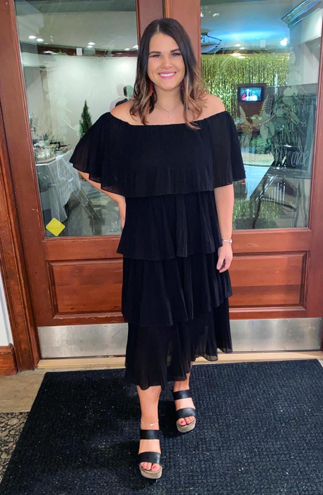 A reviewer in a tiered off-the-shoulder black dress paired with open-toe sandals stands indoors