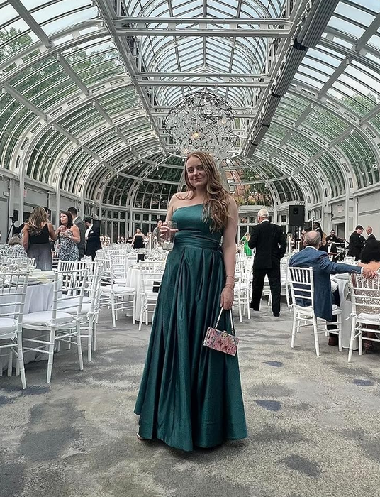 A reviewer in a sophisticated green dress stands in a venue with an arched glass ceiling and white chairs