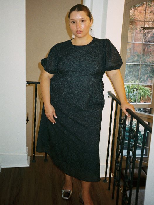 A model in a mid-length dress stands near a staircase, one hand on the railing, gazing at the camera