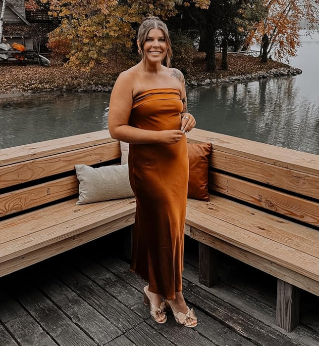 A reviewer wearing a one-shoulder dress stands on a wooden dock by a serene lake, surrounded by vibrant autumn foliage.