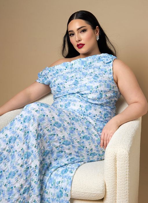 A model in a floral one-shoulder dress sits gracefully on a chair, showcasing a stylish look for a shopping feature
