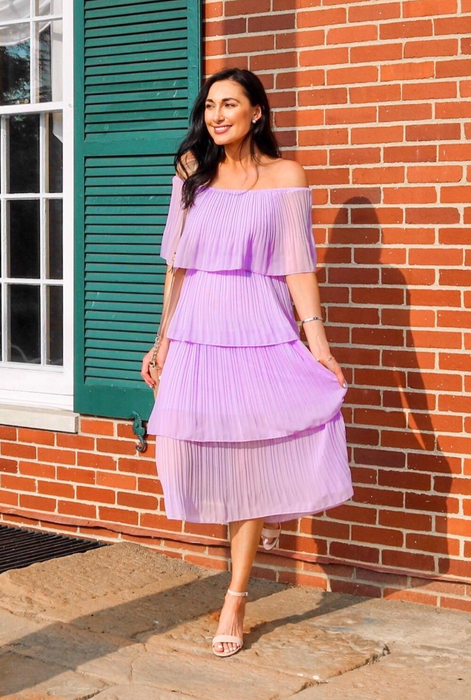 A reviewer in a pleated off-the-shoulder dress and heeled sandals strikes a pose by a brick wall