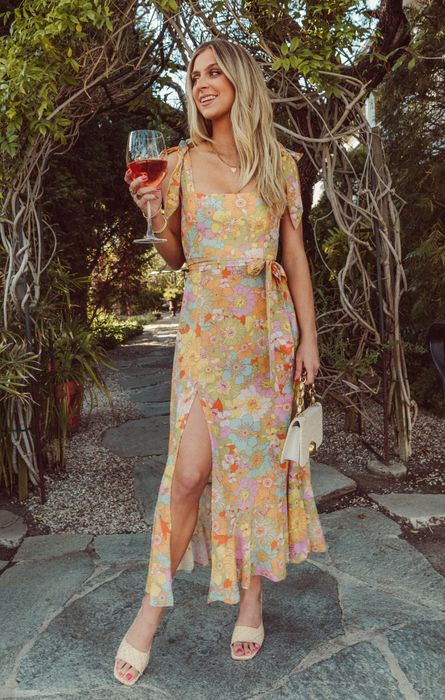 A model in a floral dress holds a wine glass and a purse while standing on a garden pathway