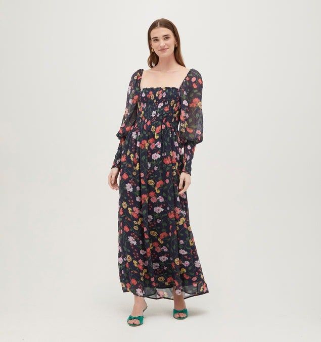 A model in a long-sleeved floral dress smiles, standing with hands slightly apart, wearing heeled sandals
