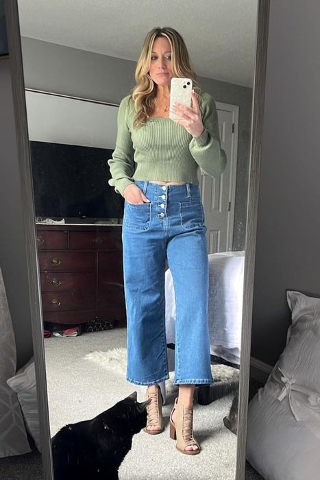 A reviewer poses for a mirror selfie in a green sweater and blue wide-leg jeans, with a black cat curiously appearing in the frame.