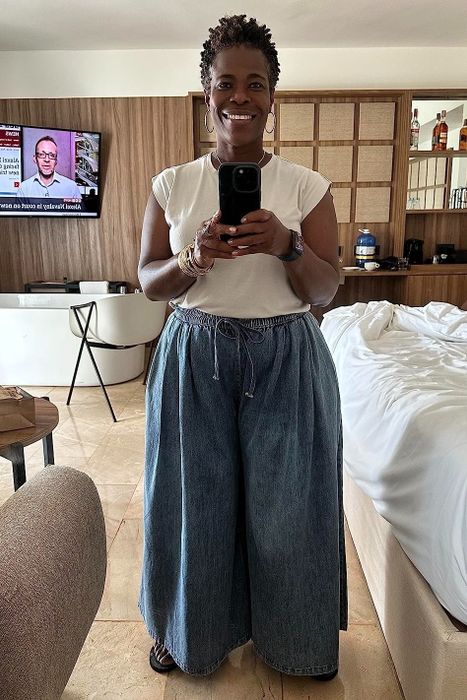 A woman in a white top and blue wide-leg pants takes a mirror selfie in a hotel room, with a table, chairs, bed, and TV visible in the background.
