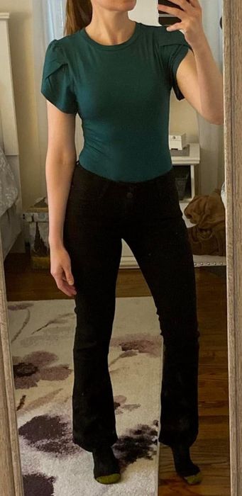 A woman dressed in a fitted short-sleeve top and high-waisted pants takes a mirror selfie in a cozy bedroom.