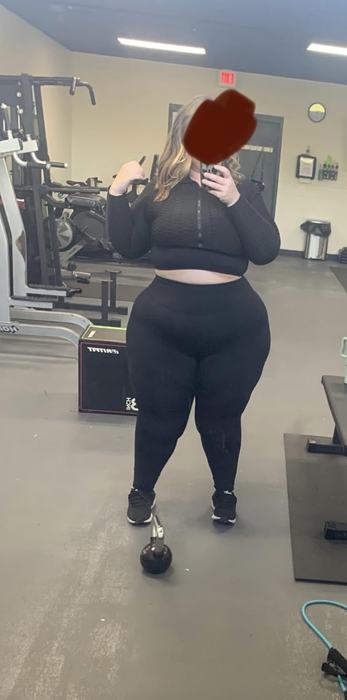 A person in a gym taking a mirror selfie, dressed in a black workout outfit with gym equipment visible in the background