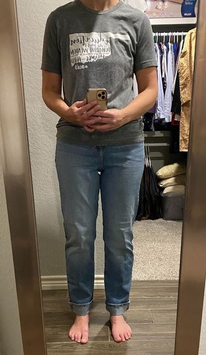 A person wearing a graphic tee and cuffed jeans, standing barefoot in front of a mirror in a room filled with clothes in the background.
