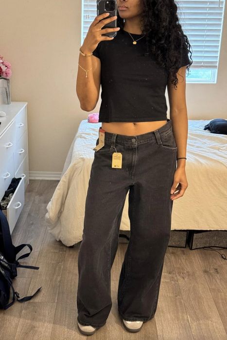 A person in a black top and wide-leg jeans snapping a mirror selfie in their bedroom.