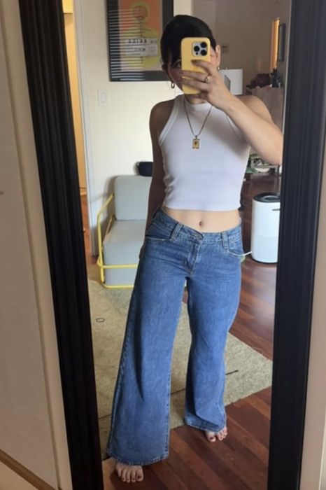 Someone in a white tank top and blue jeans taking a mirror selfie with their phone covering their face.