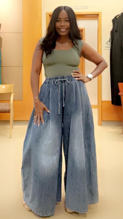 A woman stands indoors wearing a green top paired with wide-leg denim trousers, completing the casual look.