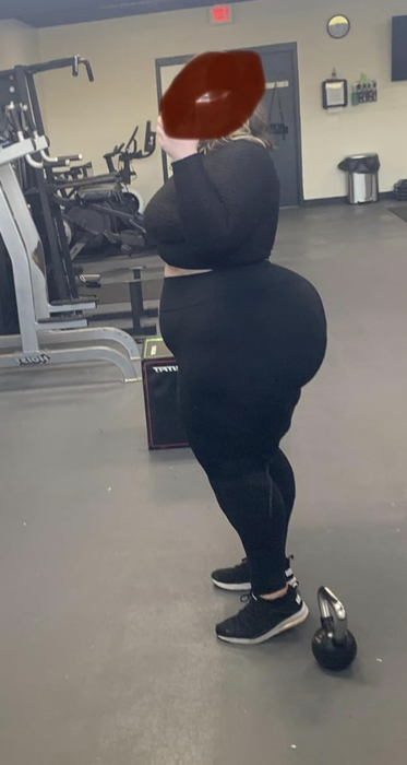 A person in gym attire standing next to a kettlebell in a workout area with exercise equipment in the background