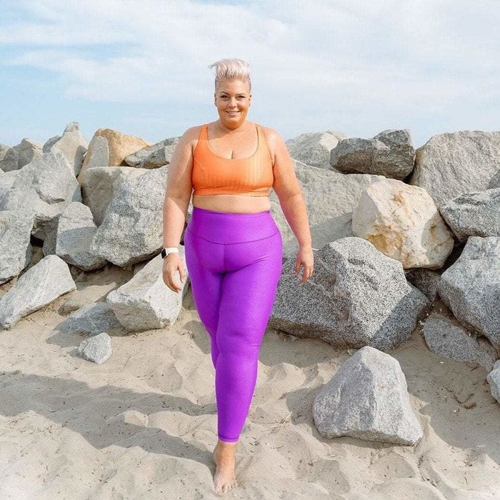 A model poses on a beach with rocks in the background, dressed in fuchsia leggings and an orange sports bra