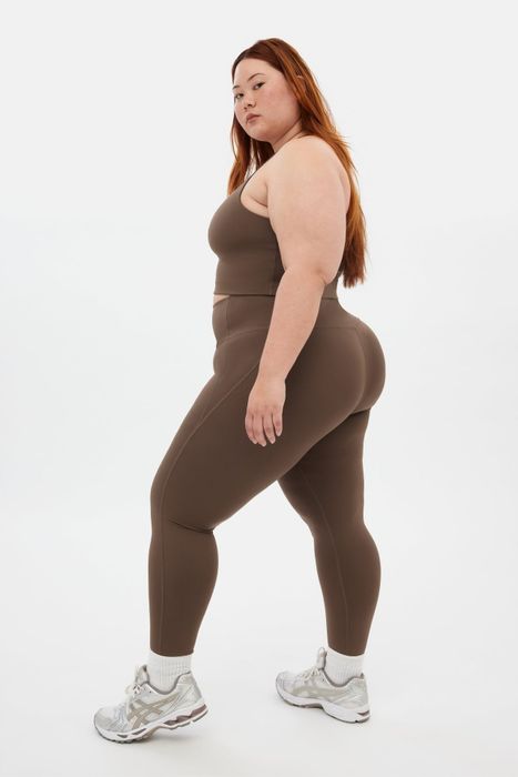 A person dressed in workout gear, including a sports bra, leggings, and athletic shoes, confidently poses in a studio environment
