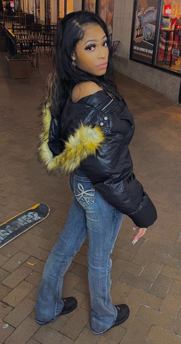 A reviewer outdoors, sporting a fur-lined black jacket and flared jeans with rhinestone accents on the back pockets.