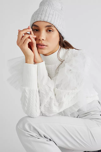 A model crouches in the front view, wearing a white knit turtleneck with tulle detailing, paired with white jeans and a white hat.