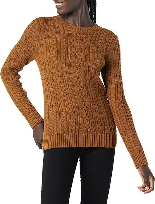 A model wears a tan fisherman cable knit sweater with blue jeans.