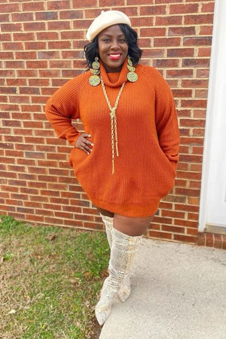 a reviewer modeling the sweater dress in orange, styled with thigh-high boots