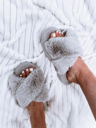 A reviewer photo of feet wearing light gray faux fur slippers with open toes and plush criss-cross straps