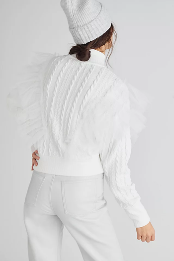 A model is seen from the back, sporting a white knit turtleneck with tulle detailing, white jeans, and a white hat.