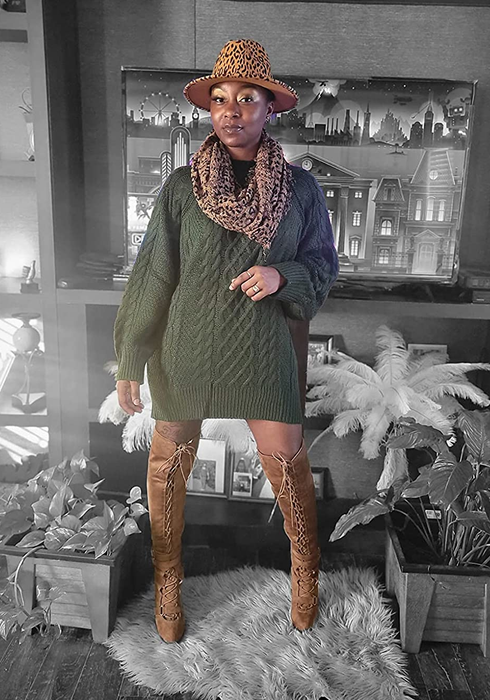 A reviewer shares a photo of themselves in a green sweater vest, brown boots, a brown scarf, and a hat.
