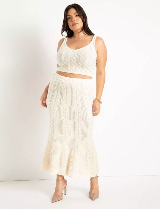 A model poses in a matching white cable-knit tank top paired with a midi skirt and nude heels.
