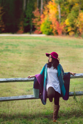 A reviewer image of a person leaning against a fence, wearing a hat, leggings, a long dress, and the purple and teal version of the shawl