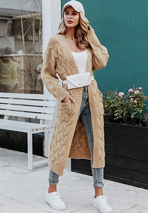 A model is styled in a long tan knit cardigan layered over a white shirt, paired with blue jeans and white sneakers.