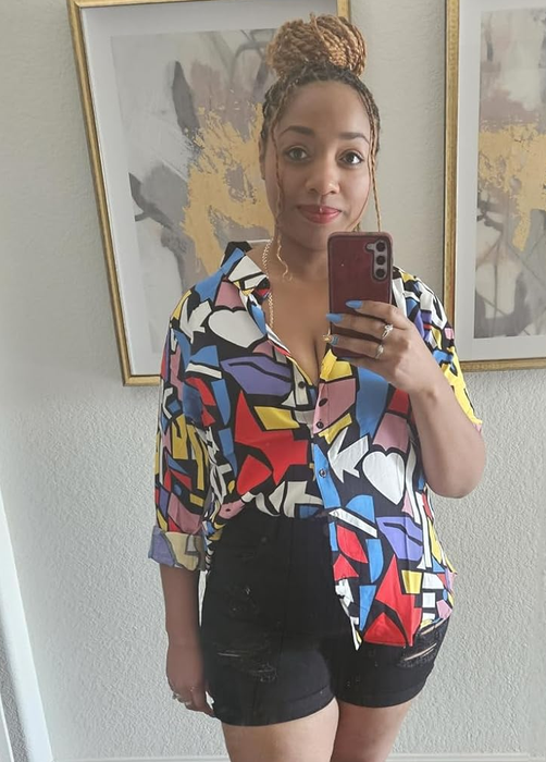 A person in a bright patterned shirt, black shorts, and yellow sandals capturing a mirror selfie with a phone