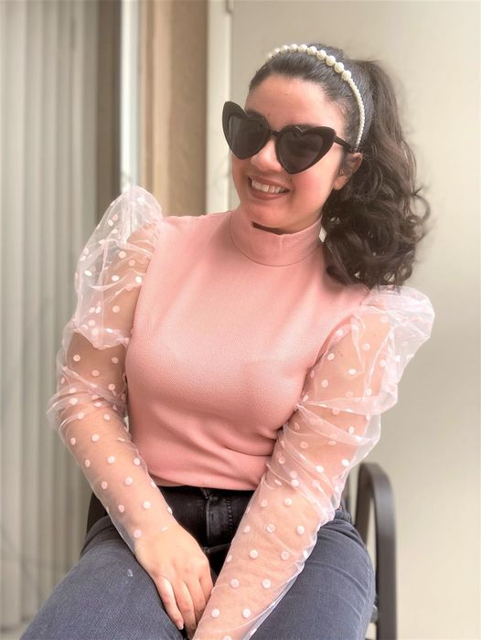 A woman wearing a pink top with sheer polka-dot sleeves and heart-shaped sunglasses, sitting and smiling