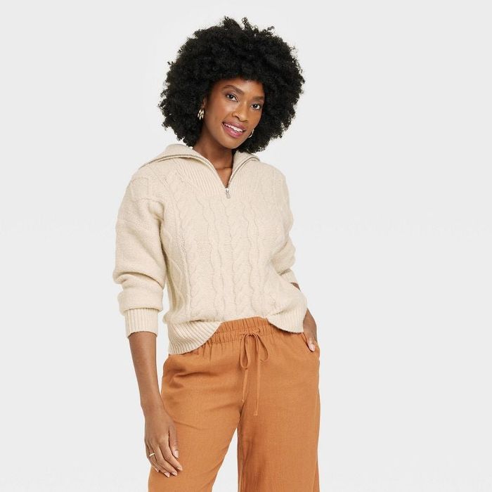 A model is wearing a cream-colored cable knit quarter zip sweater.