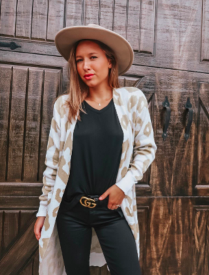 A reviewer styled in a hat, black top, dark skinny jeans, and the white and cream cheetah print duster cardigan