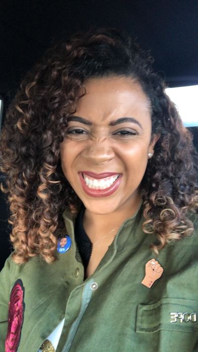 A woman with curly hair smiles broadly while wearing a green jacket decorated with pins, including a fist symbol