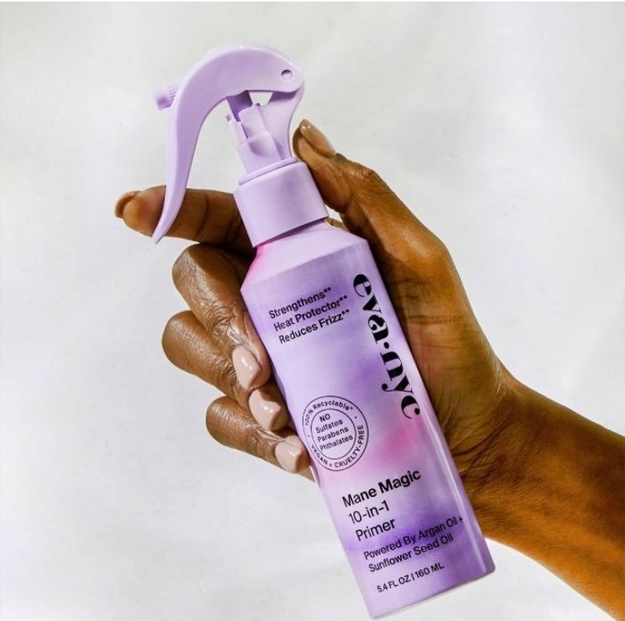 A hand holding a bottle of Eva NYC Mane Magic 10-in-1 Primer, with the label highlighting benefits like strengthening, frizz reduction, and ingredients such as argan oil and sunflower seed oil