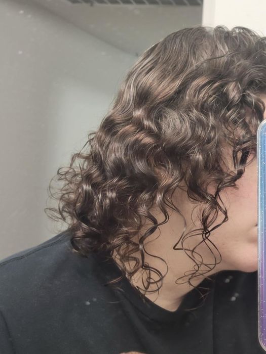 A person with curly hair takes a selfie in the mirror, showcasing their shoulder-length wavy hairstyle