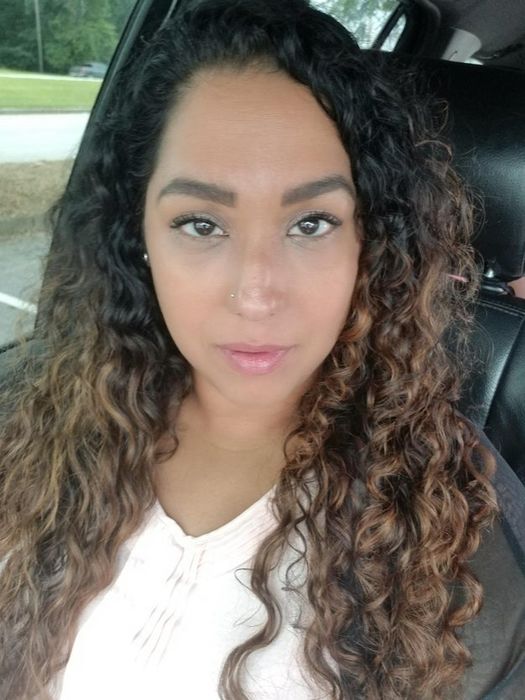 A person with long, curly hair gazes directly into the camera while seated in a car. Their expression is neutral, showing no strong emotion