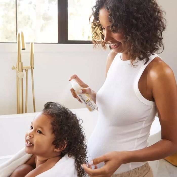 A model in a white tank top applying a hair product to a smiling child wrapped in a towel, set in a contemporary bathroom